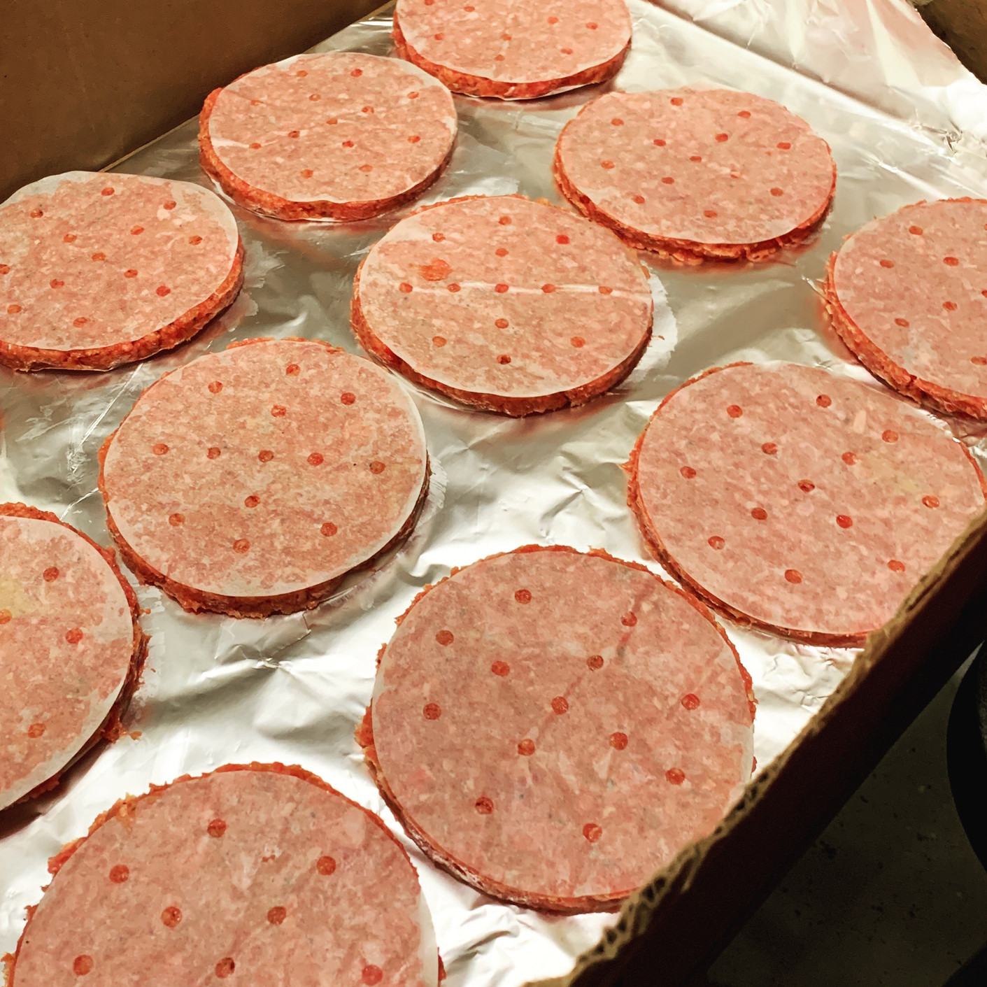 Rows of raw round meat patties with speckled texture laid on foil-lined tray, pinkish color, food prep setting