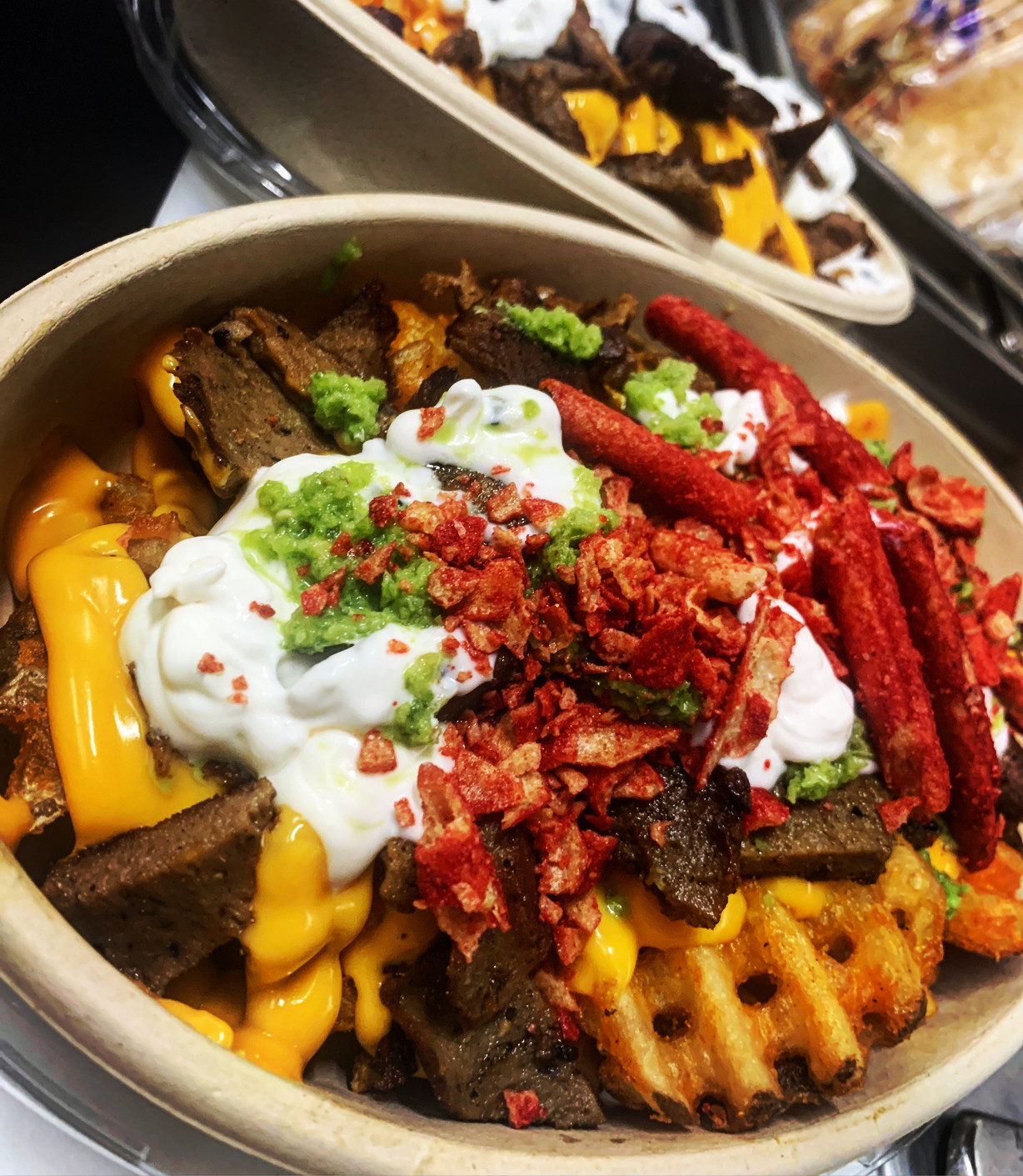 Overhead shot of a loaded bowl of waffle fries topped with chopped meat, cheese sauce, sour cream, and green sauce, warm golden tones