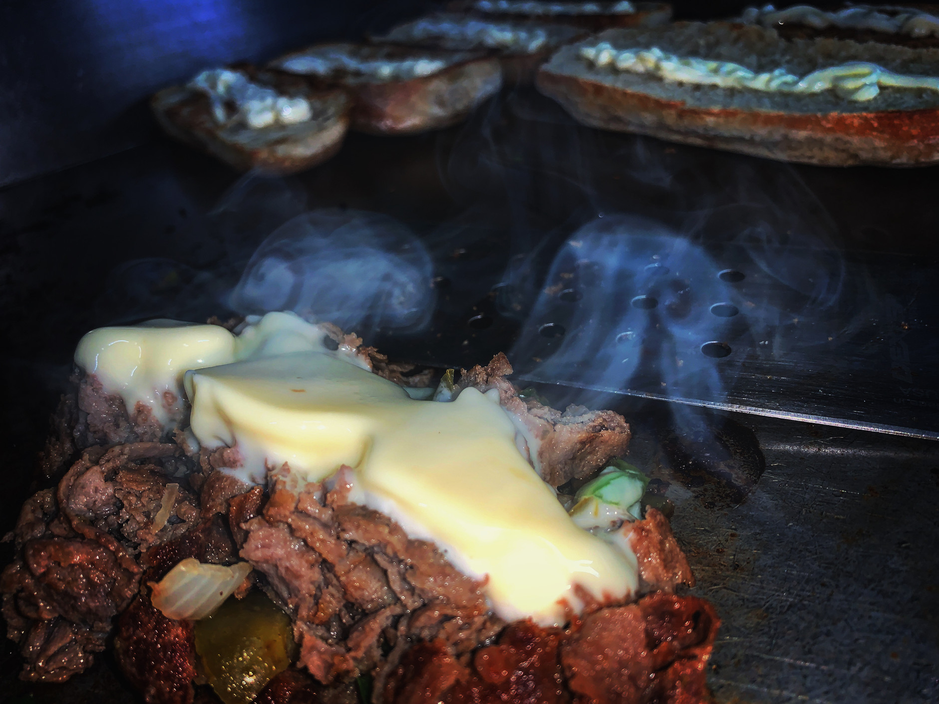 Close-up of melted cheese over grilled meat on a flattop griddle, with smoke rising and sandwich rolls in the background