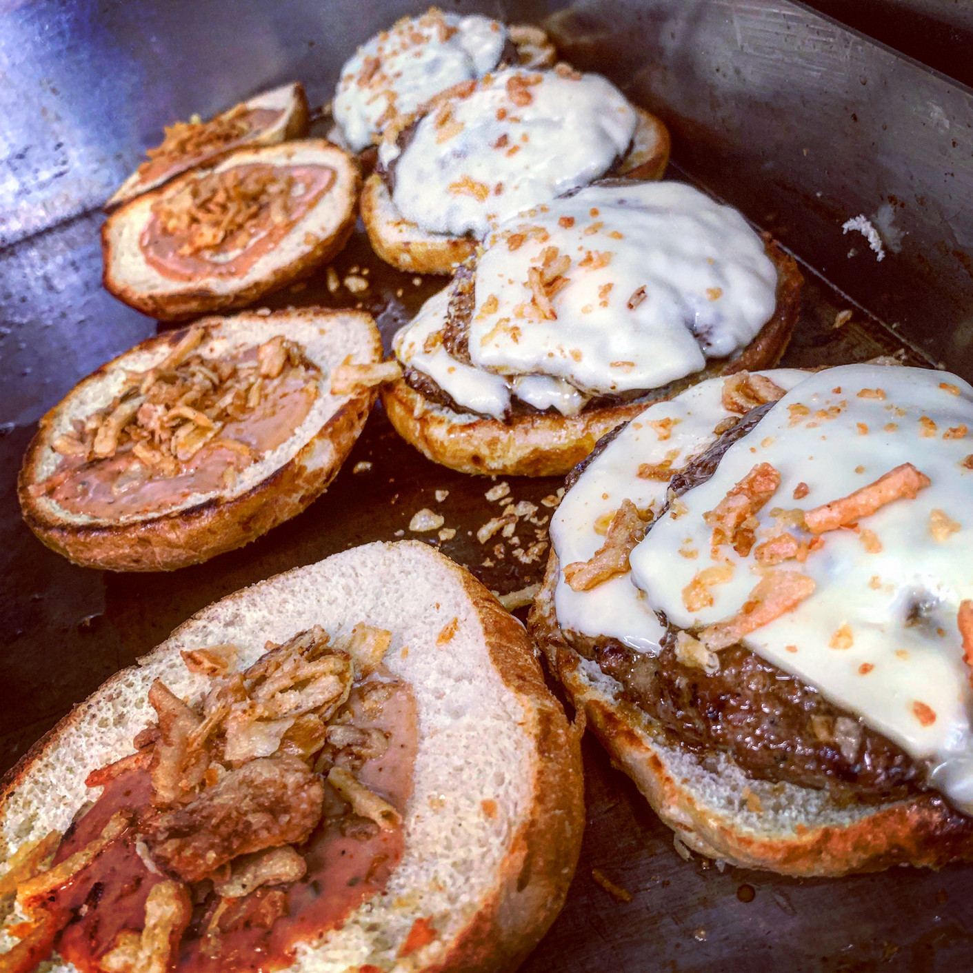 Close-up of burger patties topped with melted white cheese and crispy fried onions, on toasted buns on a metal griddle